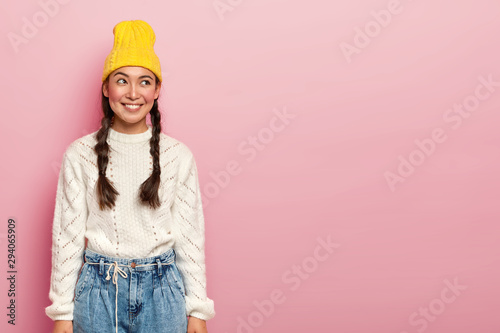 Portrait of happy Asian girl looks gladfully aside, has pleasant smile, dressed in yellow headgear, white sweater and jeans, has rouge cheeks and makeup, poses over rosy wall, free space on right side