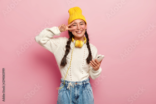 Smiling young korean woman makes peace gesture over eye, holds modern cell phone, has two plaits, smiles gently, wears yellow hat and jeans, poses against pink background. People and technology