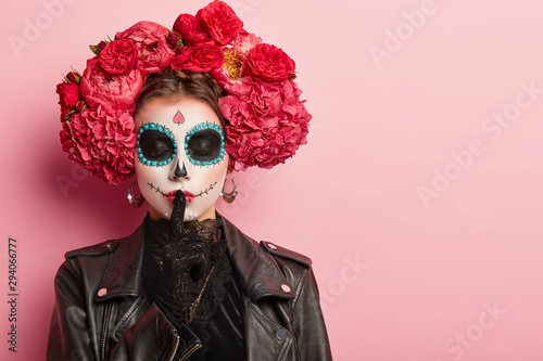 Horizontal shot of female with creative makeup applies by professional artist, dressed in black outfit, shows silence hand gesture, keeps eyes shut, poses against pink wall. Woman vampire or ghost