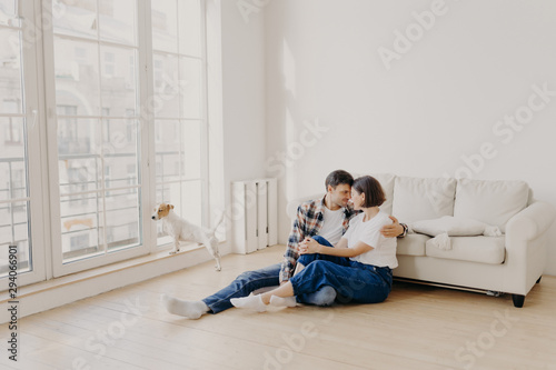 Horizontal view of happy affectionate family couple dressed in casual wear, embrace and express love to each other, pose on floor near sofa in modern apartment, their pet looks through big window