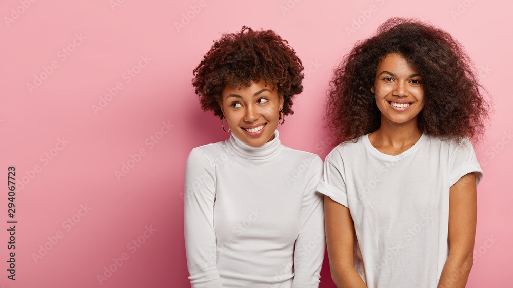 Portrait of two Afro woman with curly hair, being in high spirit, smile pleasantly, stand next to each other, wear white t shirt, turtleneck, look alike, expresses good emotions. Ethnicity, friendship - obrazy, fototapety, plakaty 