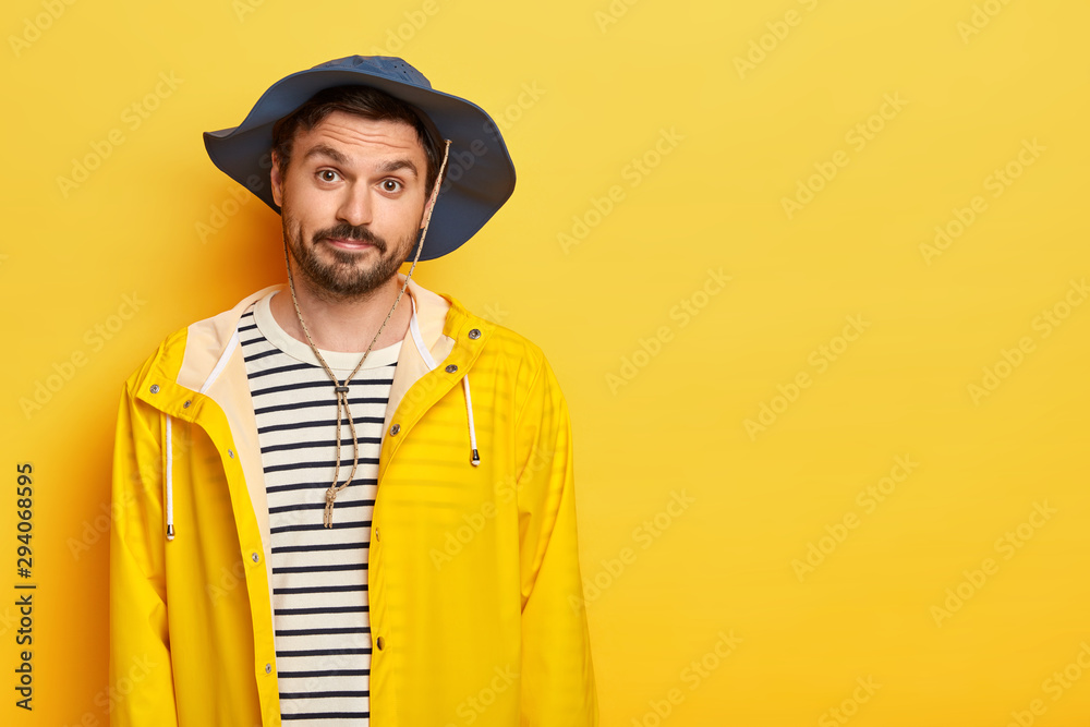 Photo of unshaven man has recreation time, going to fishing during weekend, wears hat and protective raincoat, looks with little surprisement, poses over yellow wall, free space for your text Photo of unshaven man has recreation time, going to fishing during weekend, wears hat and protective raincoat, looks with little surprisement, poses over yellow wall, free space for your text