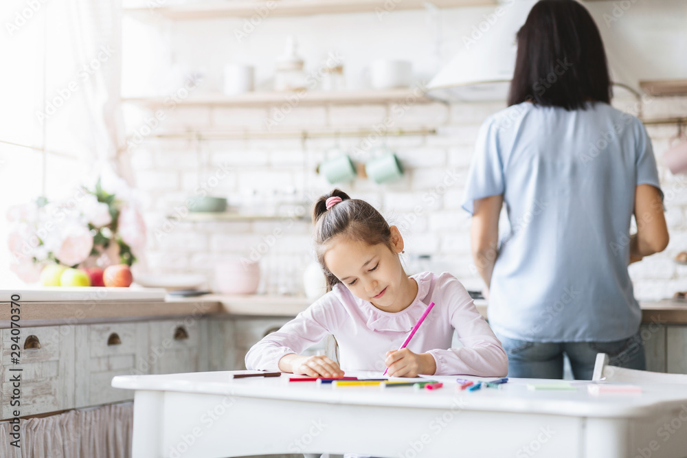 Schoolgirl doing homework in kitchen while her mother cooking food