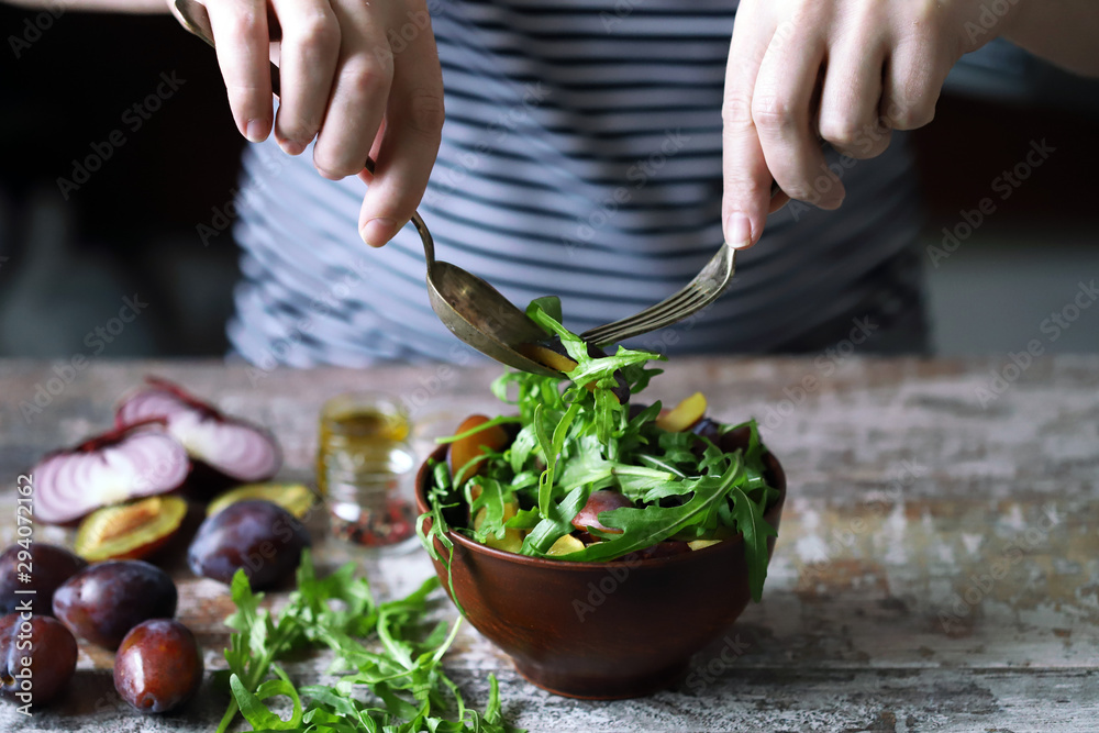 Male hands mix salad in a bowl with spoon and fork. Light salad with arugula and plums. Autumn plum salad. Healthy food. Selective focus. Macro. Male hands mix salad in a bowl with spoon and fork. Light salad with arugula and plums. Autumn plum salad. Healthy food. Selective focus. Macro.