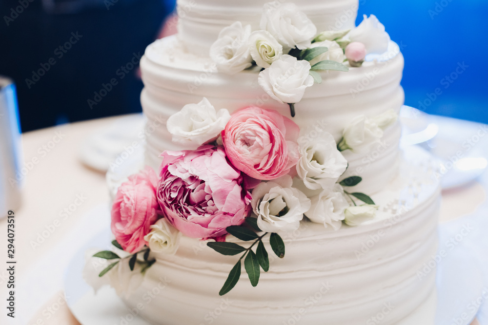 Appetizing big fresh pastry cake covered by white cream icing and decorate sweet flower serving on table. Tasty wedding event delightful dessert ready for banquet at blue light illuminate background Appetizing big fresh pastry cake covered by white cream icing and decorate sweet flower serving on table. Tasty wedding event delightful dessert ready for banquet at blue light illuminate background