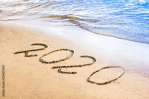 Happy New Year 2020  Text  lettering on the sand on the beach with a blue wave and sea shore  clear ocean. Numbers written on golden sunny sand. Handwritten inscription. Vacation at new year concept.