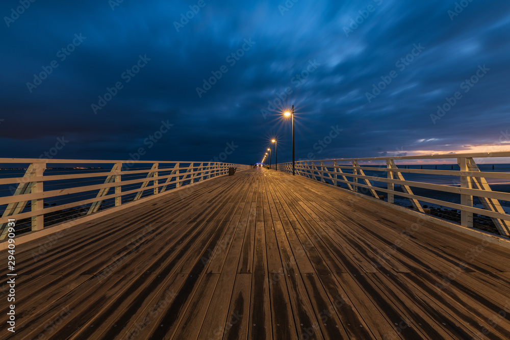 The wooden walkway with lamp street lights that stretches into the horizon on a pier during sunrise on a cloudy day in Shorncliffe, Queensland, Australia.
