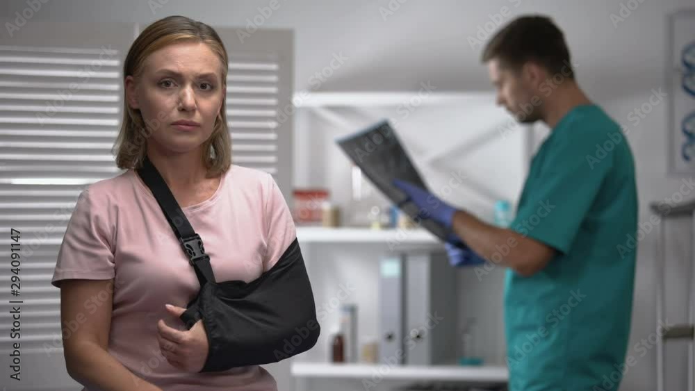 Upset female in arm sling looking at camera, radiologist checking bone x-ray Upset female in arm sling looking at camera, radiologist checking bone x-ray