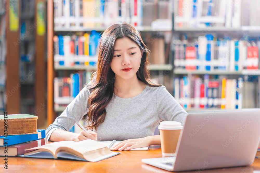 Asian young Student in casual suit doing homework and using technology laptop in library of university or colleage with various book and stationary over the book shelf background, Back to school