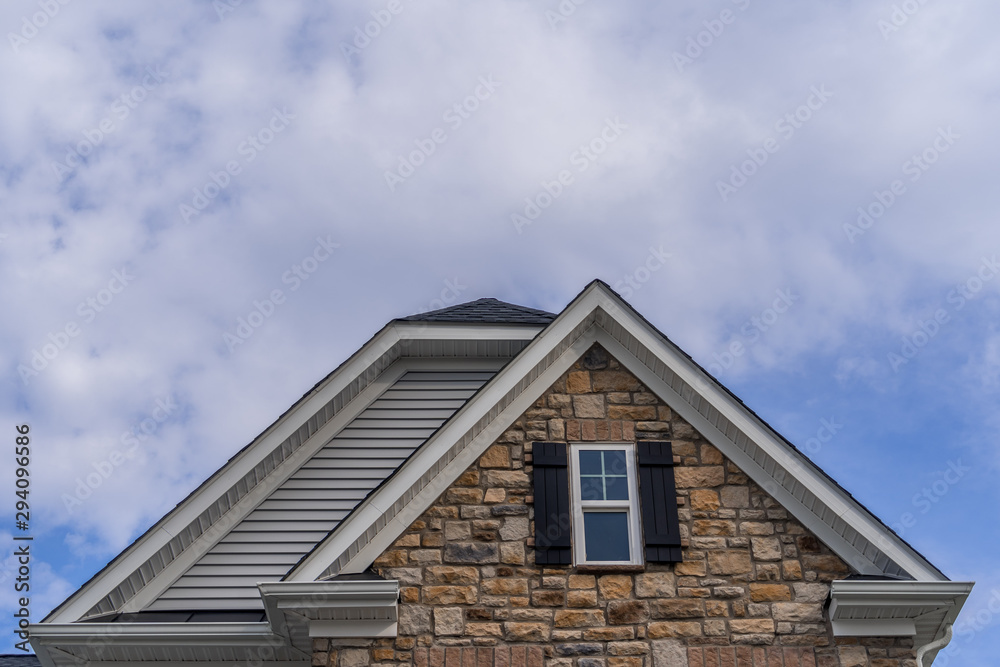 Double gable with colored stone facade siding, double hung window with white frame, vinyl shutters on a pitched roof attic at a luxury American single family home neighborhood USA