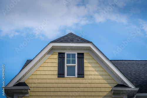 Gable with horizontal vinyl lap siding, double hung window with white frame, double vinyl shutters yellow shingle facade on a pitched roof attic at an American single family home neighborhood USA