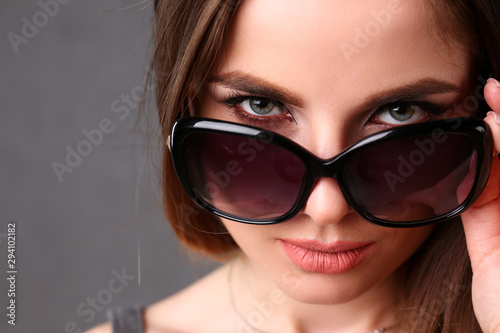 Beautiful young smiling brunette girl wearing sunglasses winking portrait over grey background. Modern sly coquette lady killer hunter plan concept