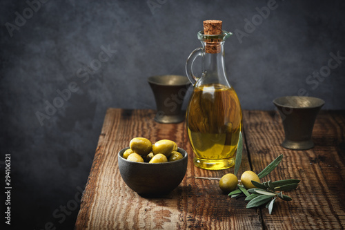Olive oil in a glass bottle, fresh green olives in a ceramic bowl and olive branch on an old wooden table.