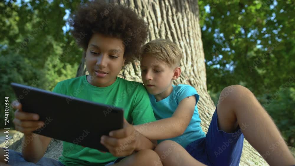 Boys playing tablet sitting under big tree in park, friends spending free time Boys playing tablet sitting under big tree in park, friends spending free time