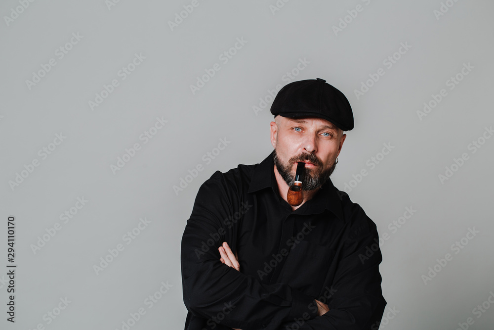 Handsome serious middle-aged man in a black shirt, hat and a pipe on gray background with copy space Handsome serious middle-aged man in a black shirt, hat and a pipe on gray background with copy space