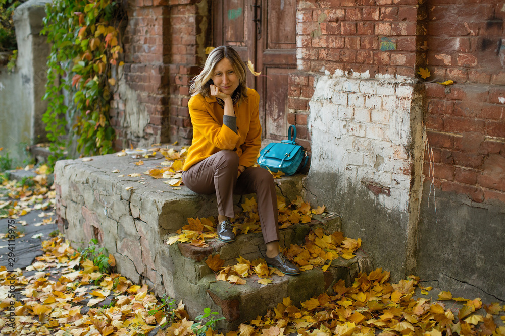 A woman in a yellow jacket and blu bag sits on the porch of an old house with maple leaves on the ground in the autumn day atmosphere.
