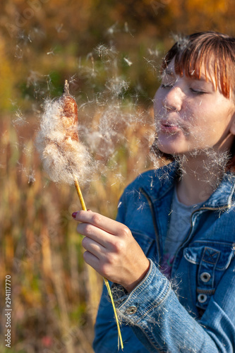 Girl child blows on a cattail in hand and fluff flies in her face. Autumn colors on a blurred background. Selective focus on the fluff.
