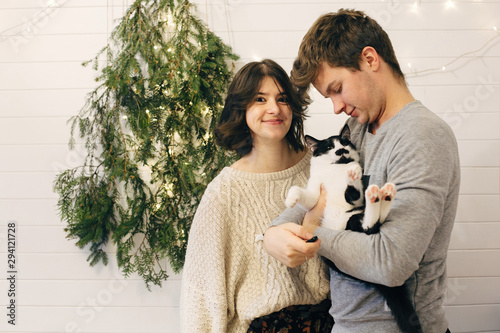 Happy hipster couple holding cat and having fun at christmas tree lights in stylish festive room. Celebrating Christmas or New Year's eve together with pets.