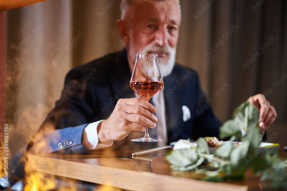 Mature beared businessman in tidy beautiful suit eat and drink champagne in romantic atmosphere. Light smoke from the fireplace on the side. Restaurant, cafe concept