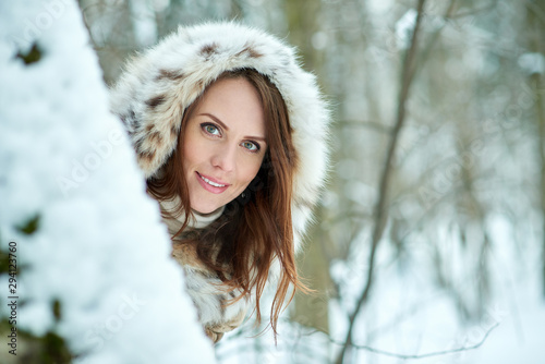 Beautiful young woman in furs looking from behind the tree on snowy forest background