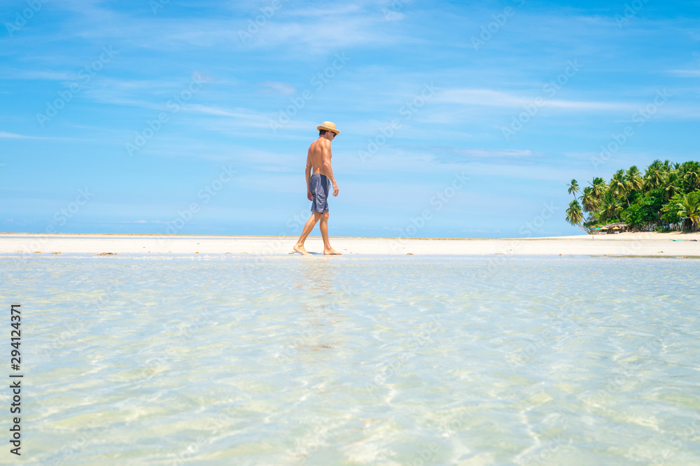 A tourist wearing a straw sun hat walking along a sand bar above the calm shallow waters of a sunny palm-lined tropical island beach 