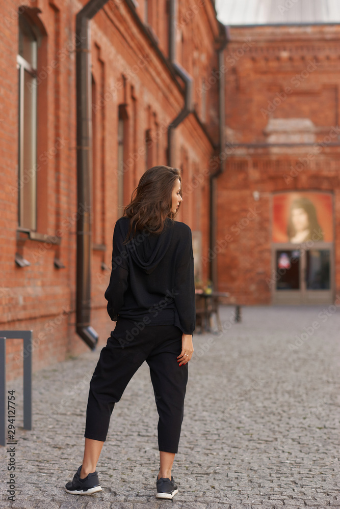 Outdoor summer portrait. Young elegant woman in black hoodie and trousers standing and posing at urban city street