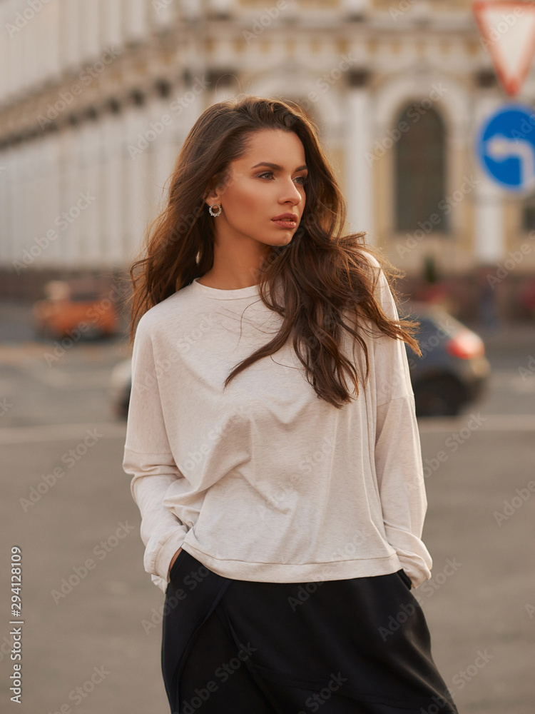 Fototapeta premium Outdoor summer portrait. Young stylish woman in white hoodie and black trousers standing and posing at city street at sunset