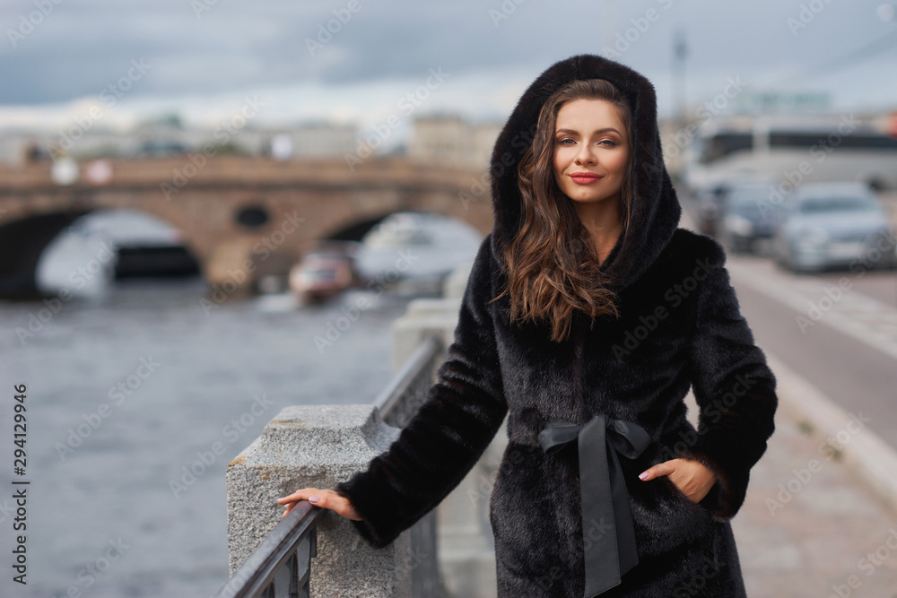 Outdoor portrait of beautiful young pretty woman in black vegan and faux fur coat standing and posing at urban city embankment. Fashion trendy style clothes