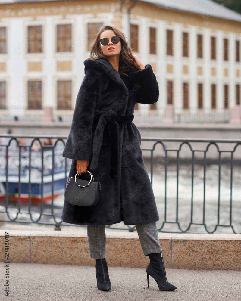 Fototapeta premium Outdoor portrait of beautiful young pretty woman in black vegan and faux fur coat standing and posing at urban city embankment. Fashion trendy style clothes