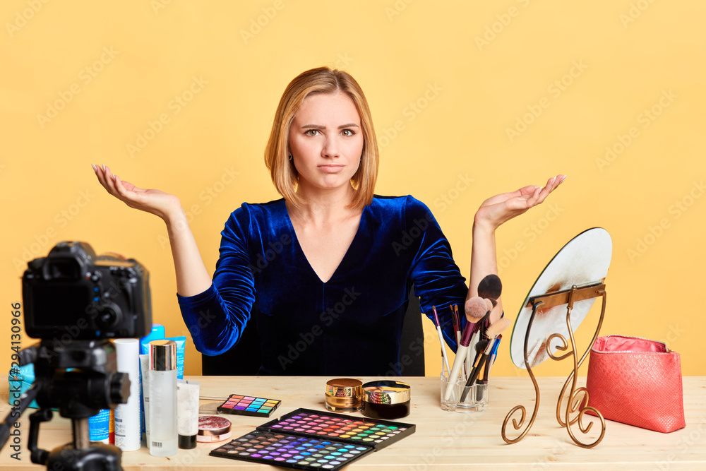 Surprised blonde female makeup artist looks with puzzlement, shruggs shoulders and spreads hands, feels clueless and uncertain, dissatisfied with something unpleasant. Emotions and massmedia concept.