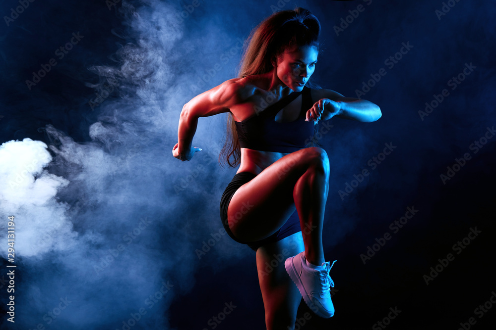 Fototapeta Young serious confident attractive sporty girl runner in shorts and top keeps her leg raised while warming up and stretching indoors. isolated black background, lifestyle