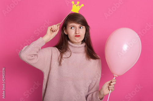 Indoor portrait of pretty brunette woman wearing funny fake crown and rose ready for party holidays time, wearing trendy cozy bright hipster sweater. Sitting on the floor with balloons