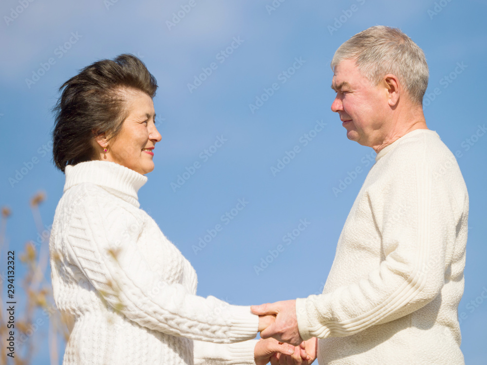 Beautiful elderly couple in white knitted sweaters close-up holding hands on a background of blue autumn sky