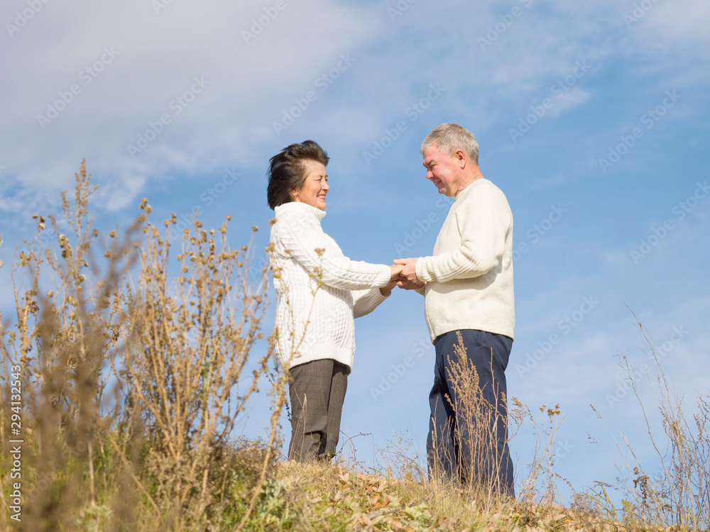 Elderly happy couple in white knitted sweaters are standing holding hands on a blue naba background in autumn afternoon. Walk the elderly. Love Elderly happy couple in white knitted sweaters are standing holding hands on a blue naba background in autumn afternoon. Walk the elderly. Love