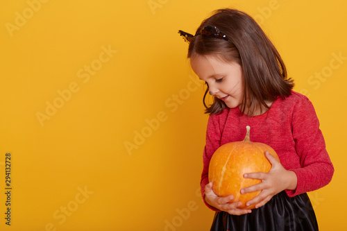 Portrait of little girl in costume of cat. Charming kid holding orange pumpkin, looking down, dresses red shirt and black skirt. Copy space for advertisment or promotion text. Halloween concept. photo