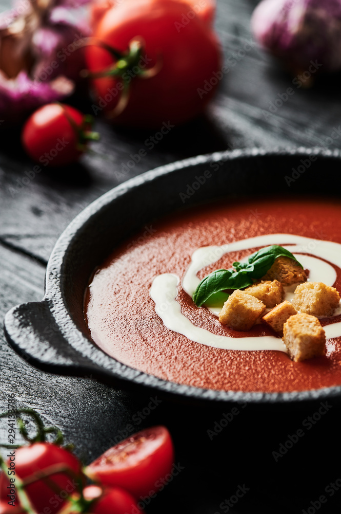 Tomato soup with cream, croutons and basil in a black bowl surrounded with tomatoes and garlic on black wooden background. Copy space for text. Selective focus. Tomato soup with cream, croutons and basil in a black bowl surrounded with tomatoes and garlic on black wooden background. Copy space for text. Selective focus.