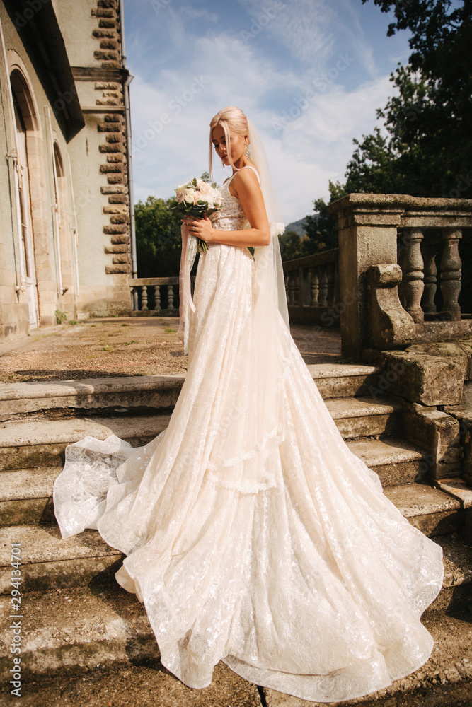 Obraz premium Gorgeous bride in wedding dress standing on the stairs by the palace. She hold a bouquet of flowers