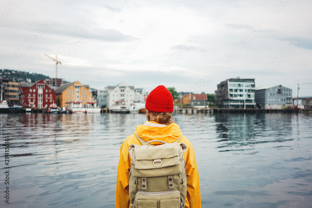 Back view of carefree man traveller with touristic rucksack wearing yellow coat and rad hat standing on berth and looking toward scandinavian city. Tourist walking the small authentic city among ship Back view of carefree man traveller with touristic rucksack wearing yellow coat and rad hat standing on berth and looking toward scandinavian city. Tourist walking the small authentic city among ship