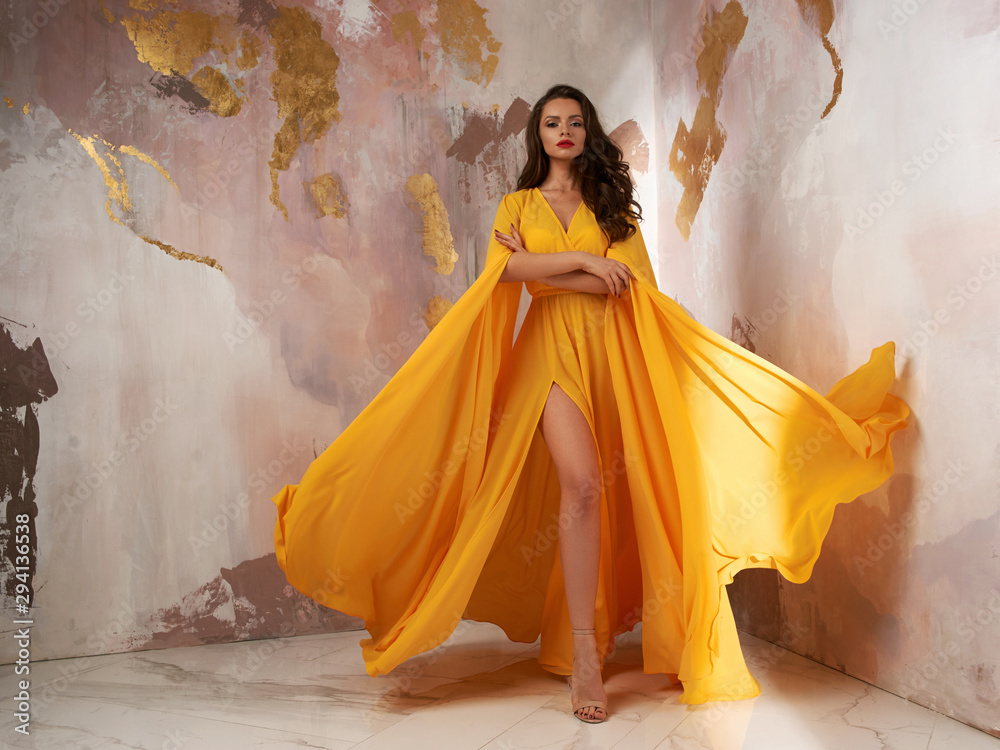 Young beautful caucasian woman with long wavy brunette hair in yellow flying dress posing against wall. Vogue style fashion portrait - obrazy, fototapety, plakaty 