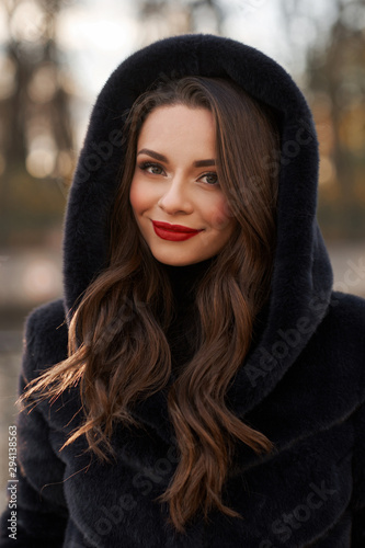 Pretty lady in black fur coat with hood standing and posing at city street. Fashionable woman. Closeup face portrait.