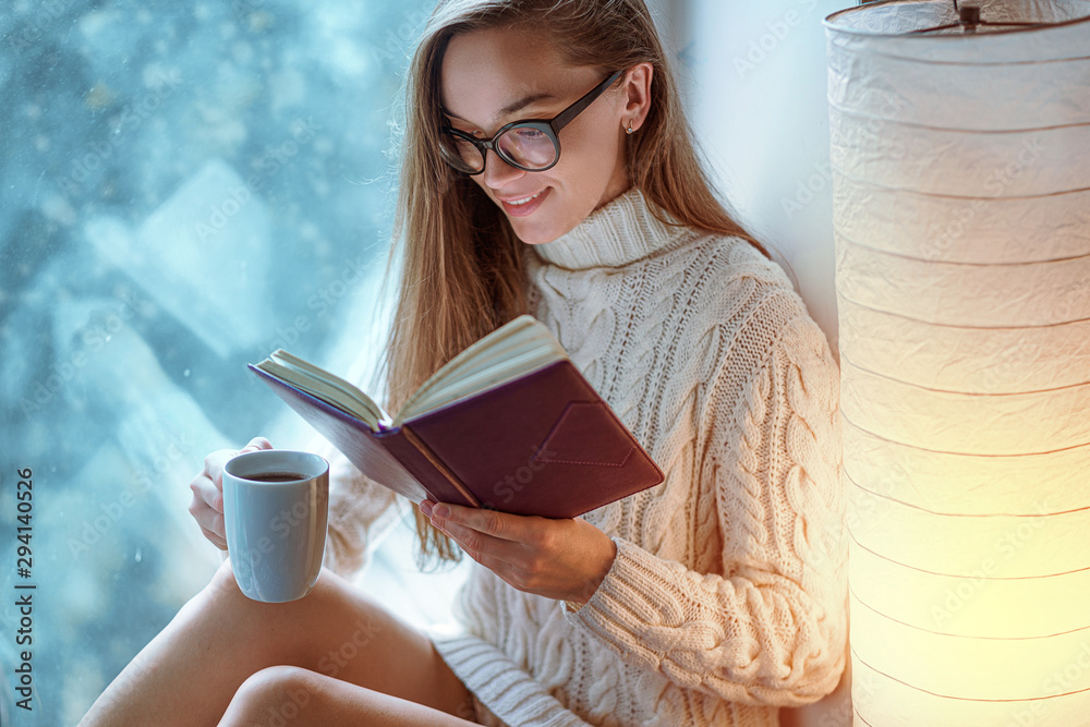 Obraz premium Portrait of comforting young happy cozy woman in glasses in knitted winter white warm sweater drinks a cup of hot cocoa during reading favorite book on a window sill by the window at home in winter