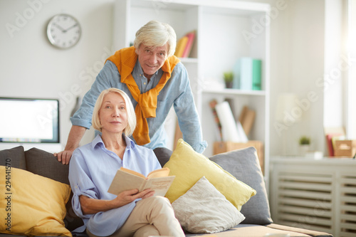 Portrait of modern senior couple in home interior focus on woman reading book sitting on comfortable couch, copy space