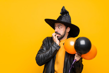 Man with witch hat holding black and orange air balloons for halloween party nervous and scared putting hands to mouth