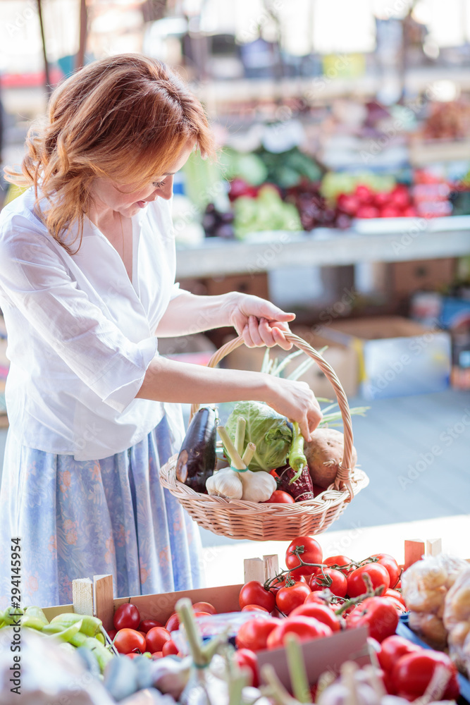 Happy mature woman buying fresh organic vegetables on a local farmer's marketplace. Holding a basket filled with various vegetables. Healthy food production and eating