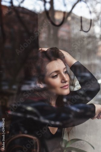 Beautiful girl with long brunette haibeside white flowers behind the glass, coffee shop window, concept of female emotions