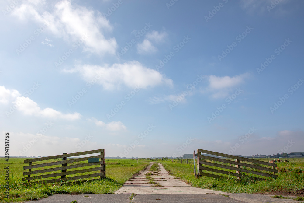 Gate that is open, wooden gate in agricultural land, bright green meadow with sky with clouds and clear horizon in the background.