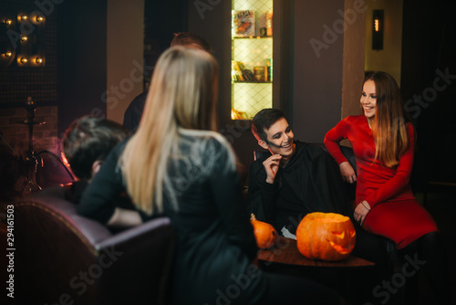 Bunch of friends from two guys and two girls having fun celebrating Halloween in a dark cozy cafe. Carved pumpkin on the table. Colorful make-up of young people.