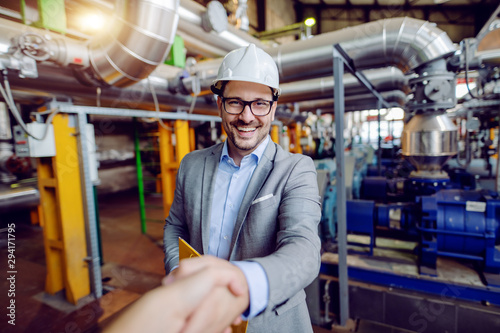 Handsome caucasian manager in suit and with helmet on head holding folder with documents and shaking hands with investor while standing in power plant.