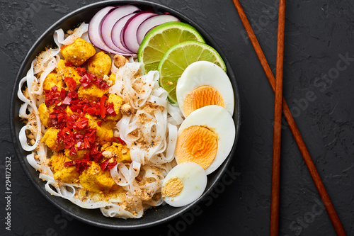 Nan Gyi Thoke in black bowl at dark slate background. Nan gyi Thohk is popular burmese cuisine dish with rice noodles, chicken breasts with spices, eggs, peanuts, lime and red onion. Top view photo
