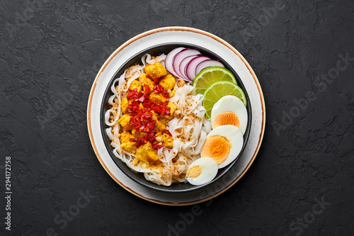 Nan Gyi Thoke in black bowl at dark slate background. Nan gyi Thohk is popular burmese cuisine dish with rice noodles, chicken breasts with spices, eggs, peanuts, lime and red onion. Top view